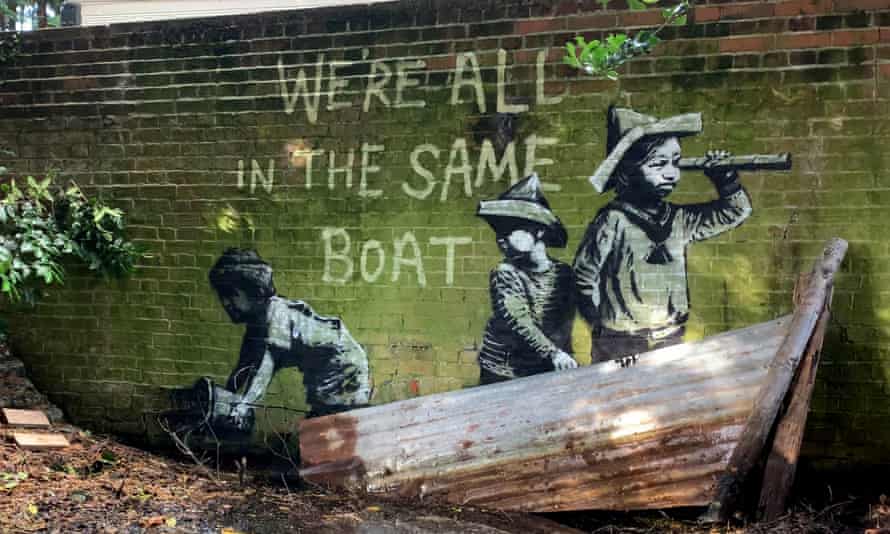 Графити на Банкси върху тухлена стена със зелена боя, три деца в лодка и текст „всички сме в една лодка“