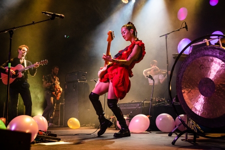 music band Japanese breakfast is performing on the stage, the front woman Michelle in her red dress and black boots is jamming her electro, colourful ballons are scatered on the stage dimly lit
