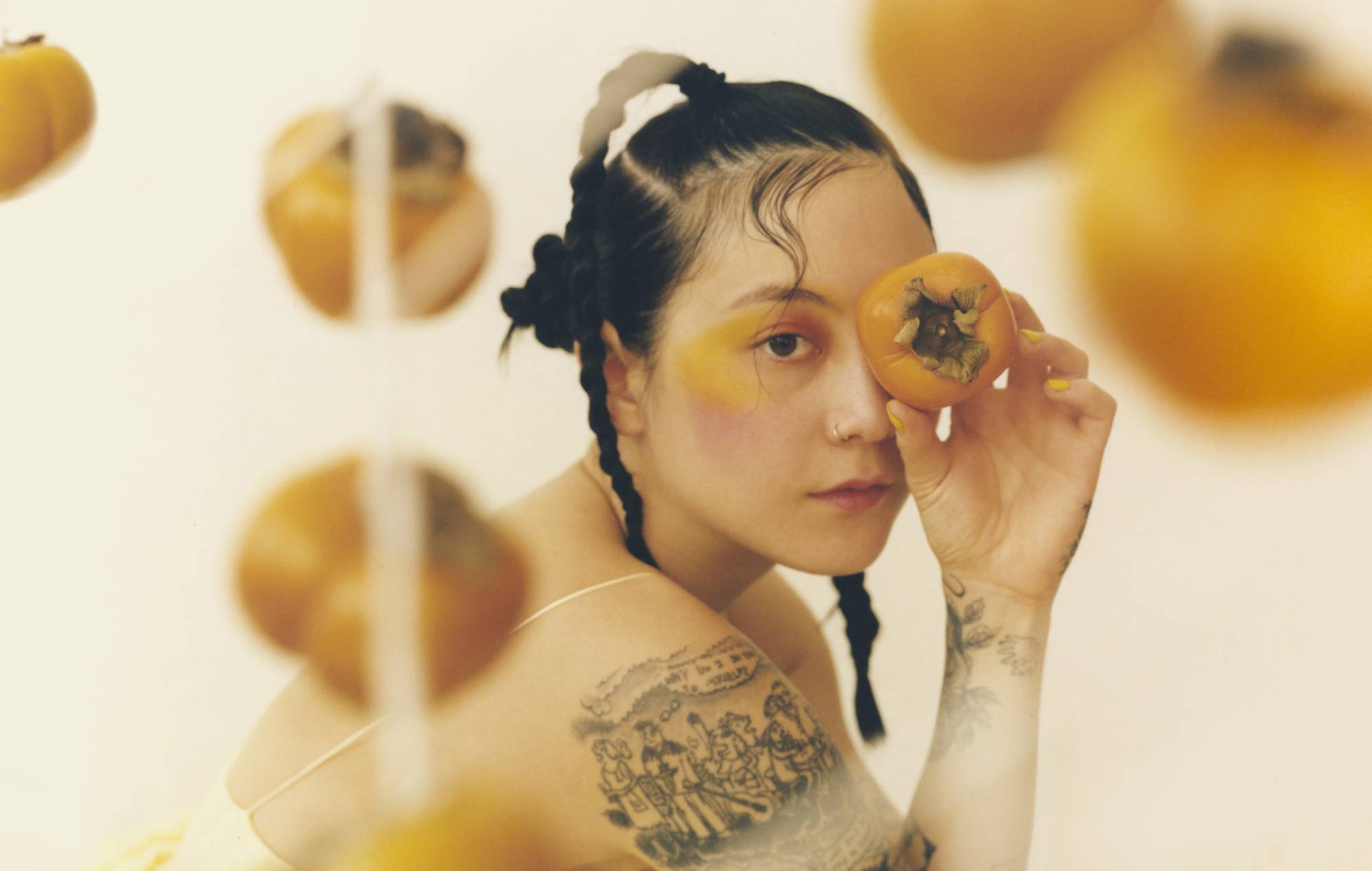 Singer Michelle Zauner squatting and covering her left eye with an orange round fruit she is holding as many other identical fruits hanging in the front