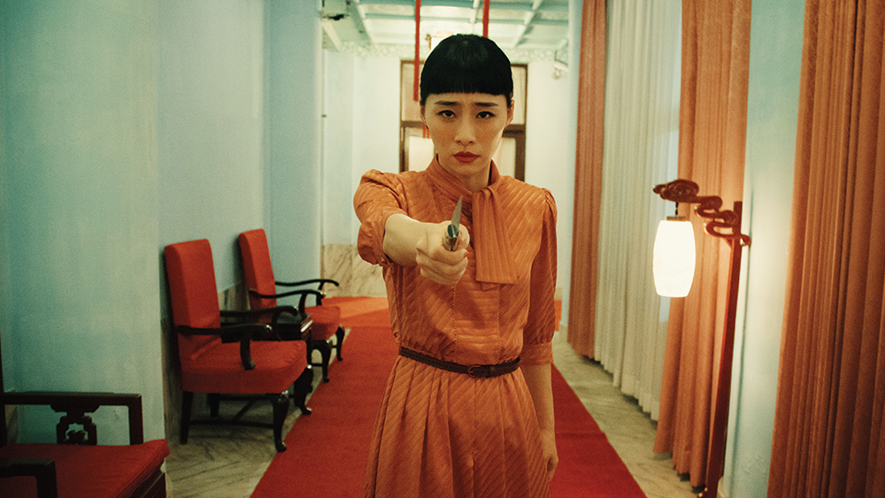 Nina Wu in her orange dress with black thin belt, holding the knife towards the camera, standing in a long corridor furnished with red cushioned sofa chairs and long cushions