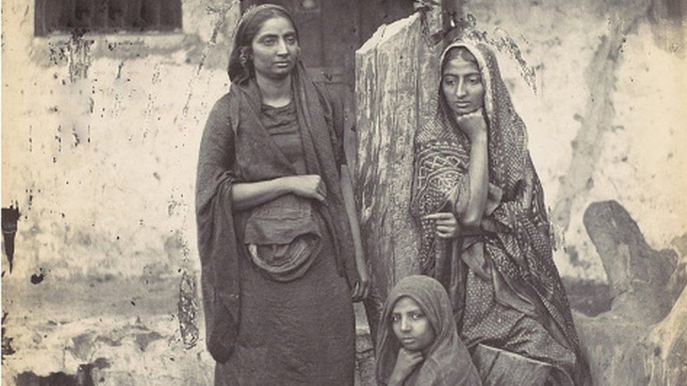 An opaque photograph of three Indian women in colonial India, standing in front of a rented home.