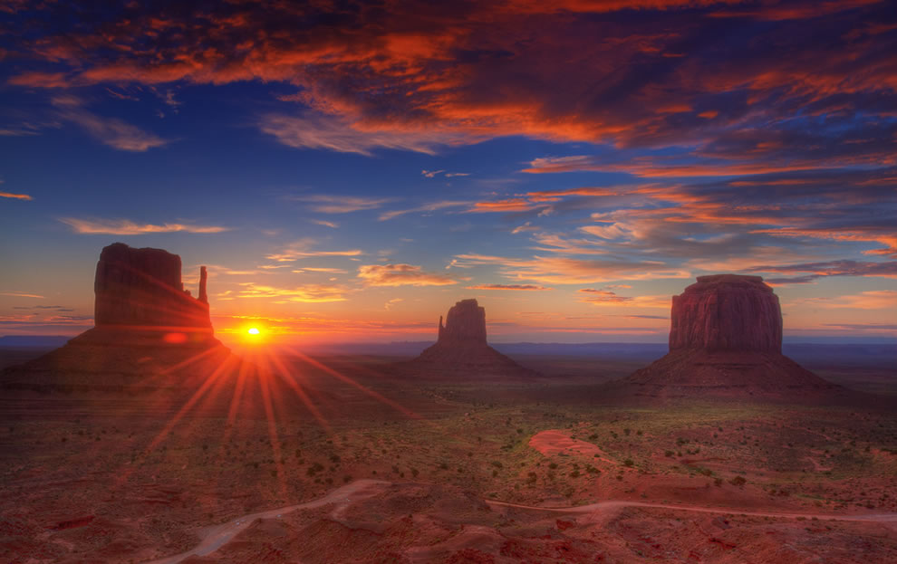 пейзажът на земите на навахо има три планини, засенчени от оранжево облачно небе, докато слънцето залязва на запад