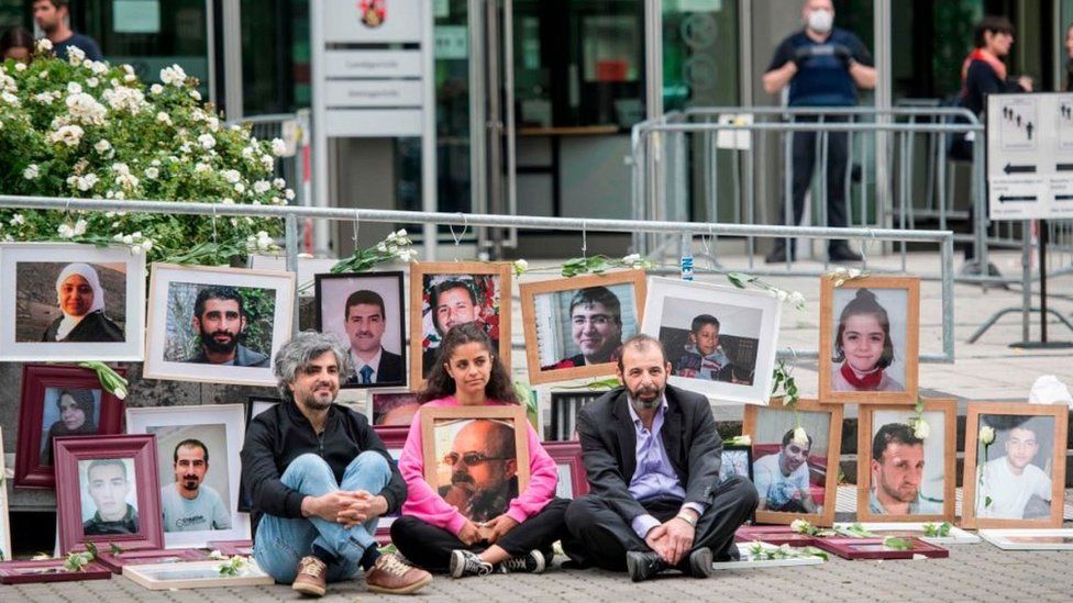 Et fotografi af syriske flygtninge og/eller ofre, der sidder uden for området omkring det tyske retshus, omgivet af fotografier af dem, der er gået tabt under borgerkrigen under de syriske soldater i interneringscentrene.