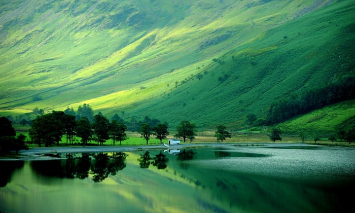 A photo of the Lake District shows high green mountains turning the tree lined lake below them into a blue-green hue.