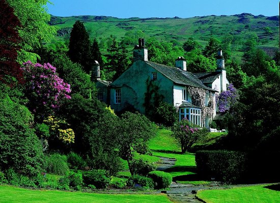 A photo of Woodsworth's idylli cottage in the Lake District nestled between two hills and surrounded by trees and gardens under a bright blue sky. 