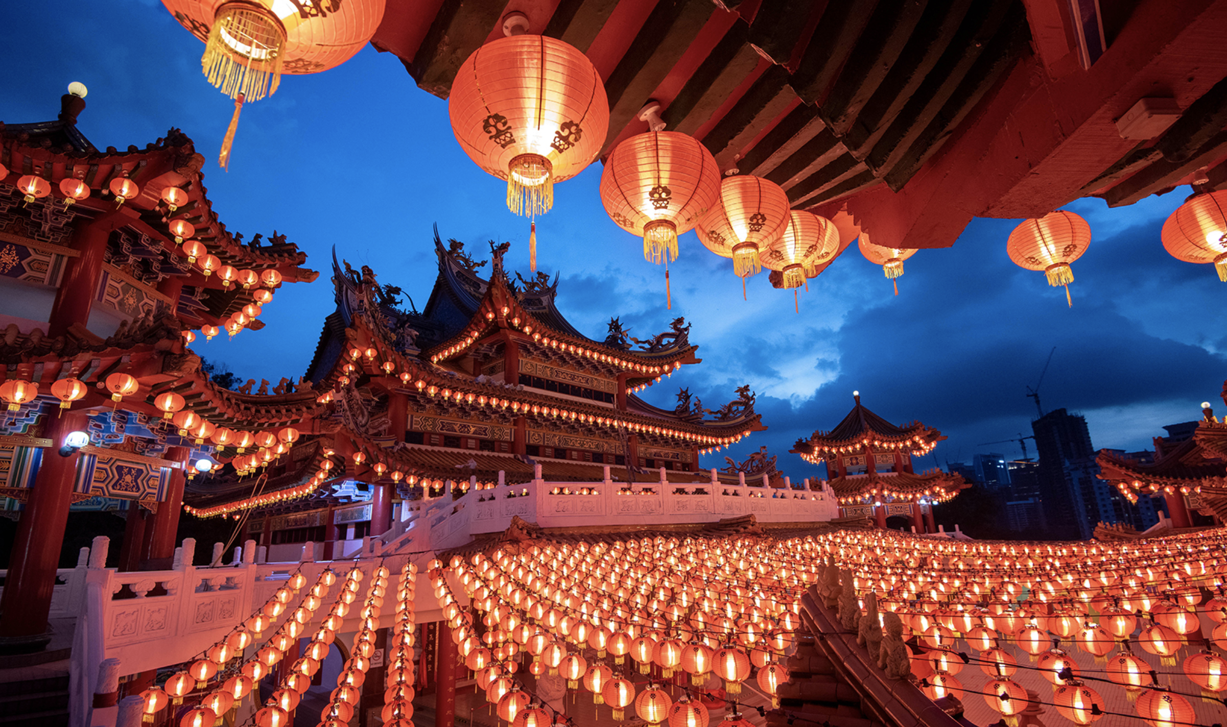 Малайзийски храм стои срещу синьо нощно небе, със светещи червени фенери в редове на пода и на преден план