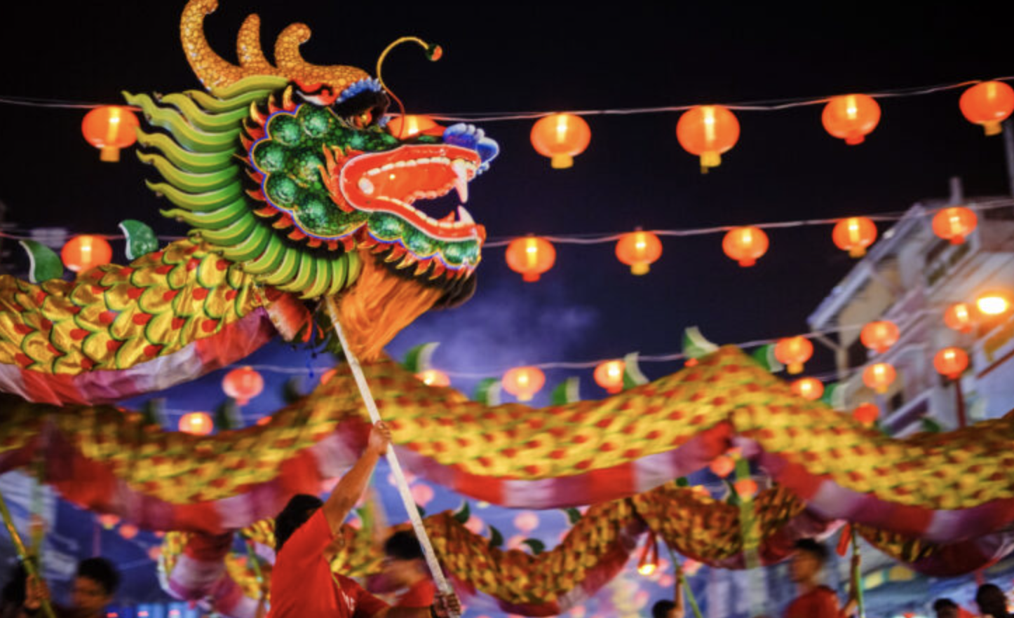 A dragon is lit up and carried around by dragon dancers in the night with red lanterns above the dragon's head in a Lunar New Year festival