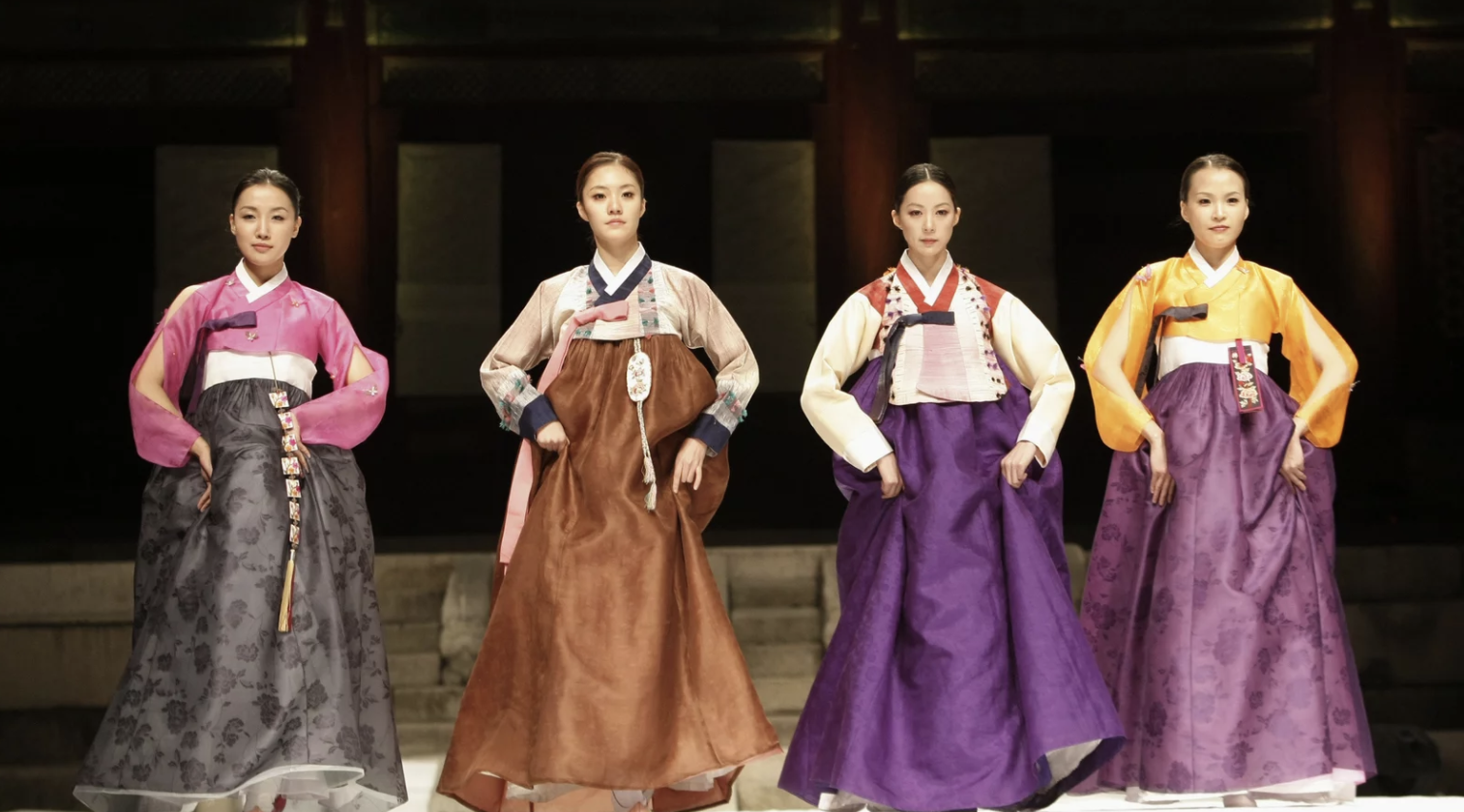 Four women wearing hanbok in different colors are shown with their hands on their hips posing in front of a dark background