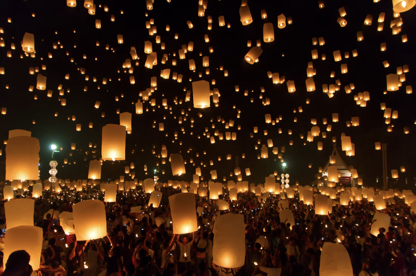 A night sky is filled with yellow lanterns floating with people lifting them off into the air