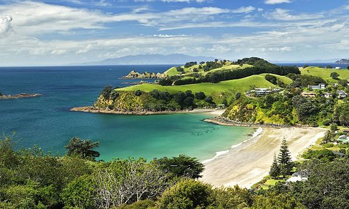 A shot of Waiheke Bay in New Zealand shows white sand, blue waters and green hills. 