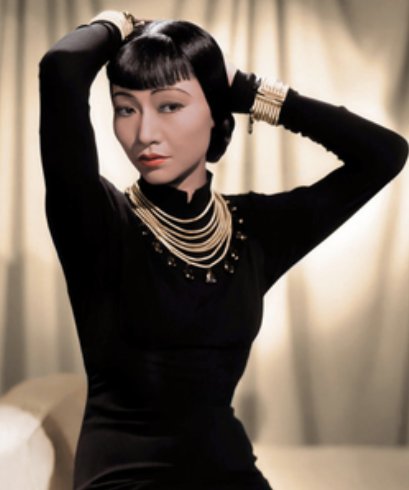 Anna May Wong is posing in a tight long sleeve black dress and gold jewelry against a white curtain for a picture