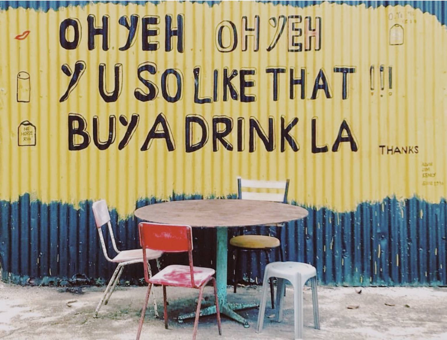 The painted words on a wall read "Oh Yeh Oh Yeh, Y U So Like That!!! Buy A Drink La" in front of a table and chairs