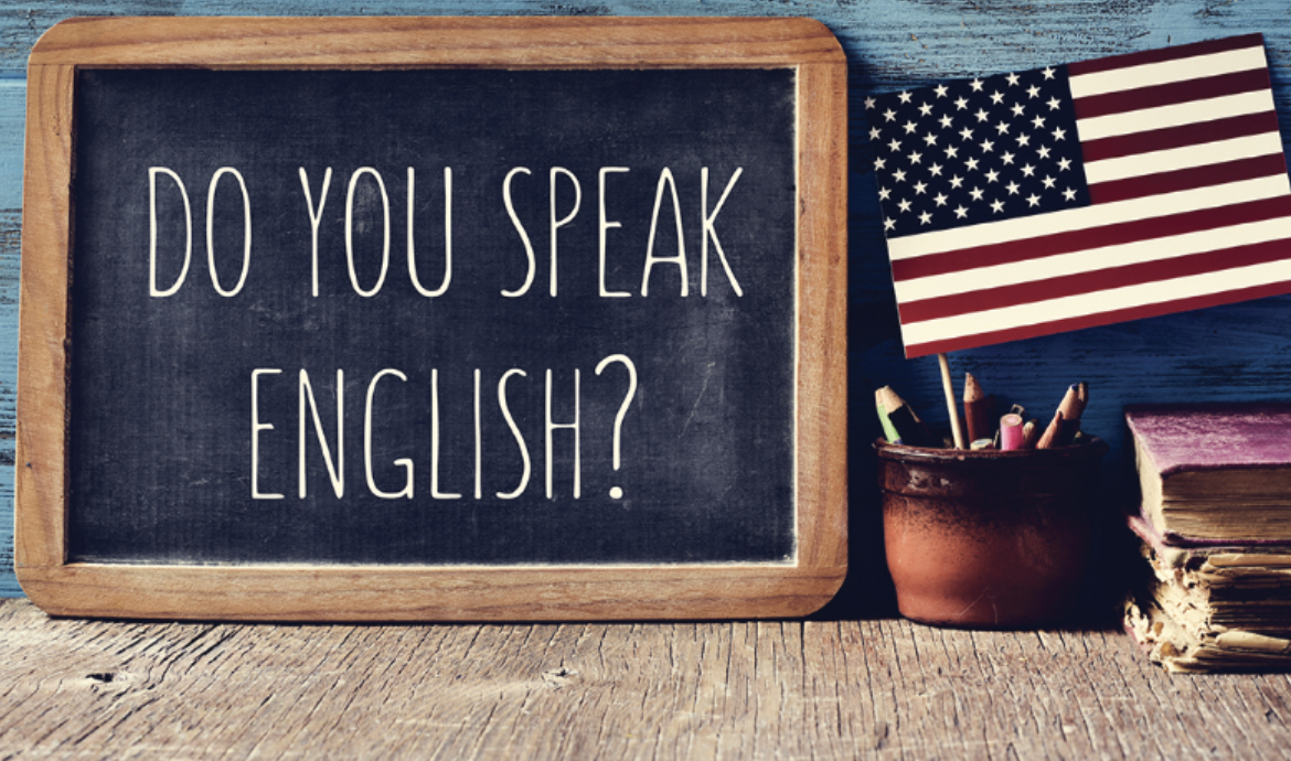 A sign that says "Do you speak English?" is next to an American flag and books on a table