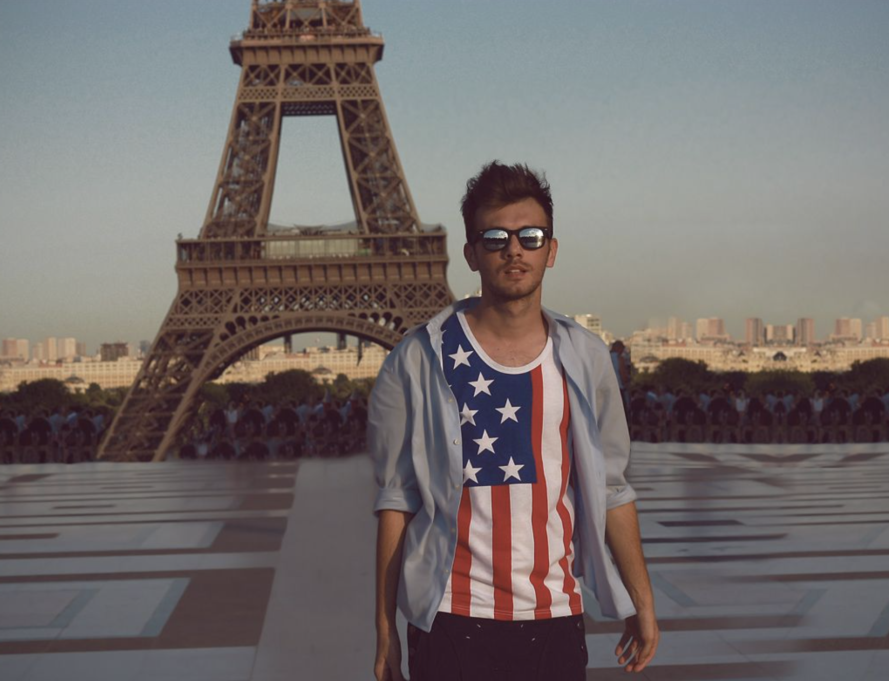 An American white man is wearing sunglasses and a shirt with the American flag in front of the Eiffel Tower