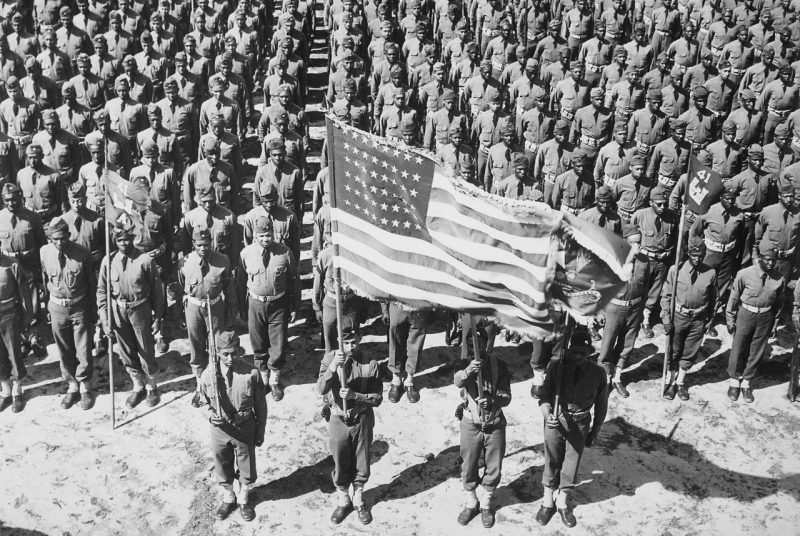 A black and white photograph of the American soldiers, lined up in uniformed order, four in the front of the many rows with the second soldier from the right holding a flowing American flag.