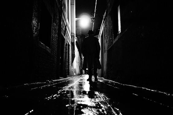 Черно-бяла снимка на мъж с тренчкот, който върви по залъскана от дъжда алея.